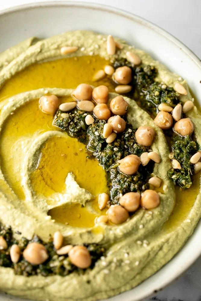 Bowl of creamy pesto hummus served with fresh vegetables and pita bread.